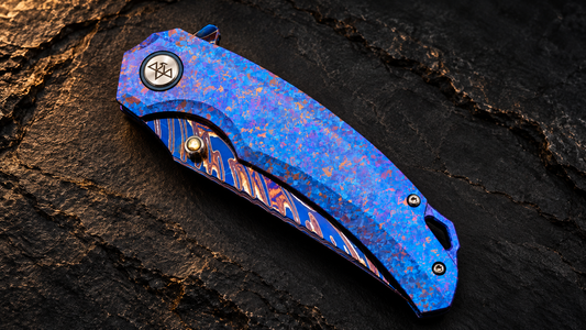 VORIXN Rimeflare KB132 folding knife with a blue-purple crystallized titanium handle and colorful Damascus blade, closed view on a dark stone background.