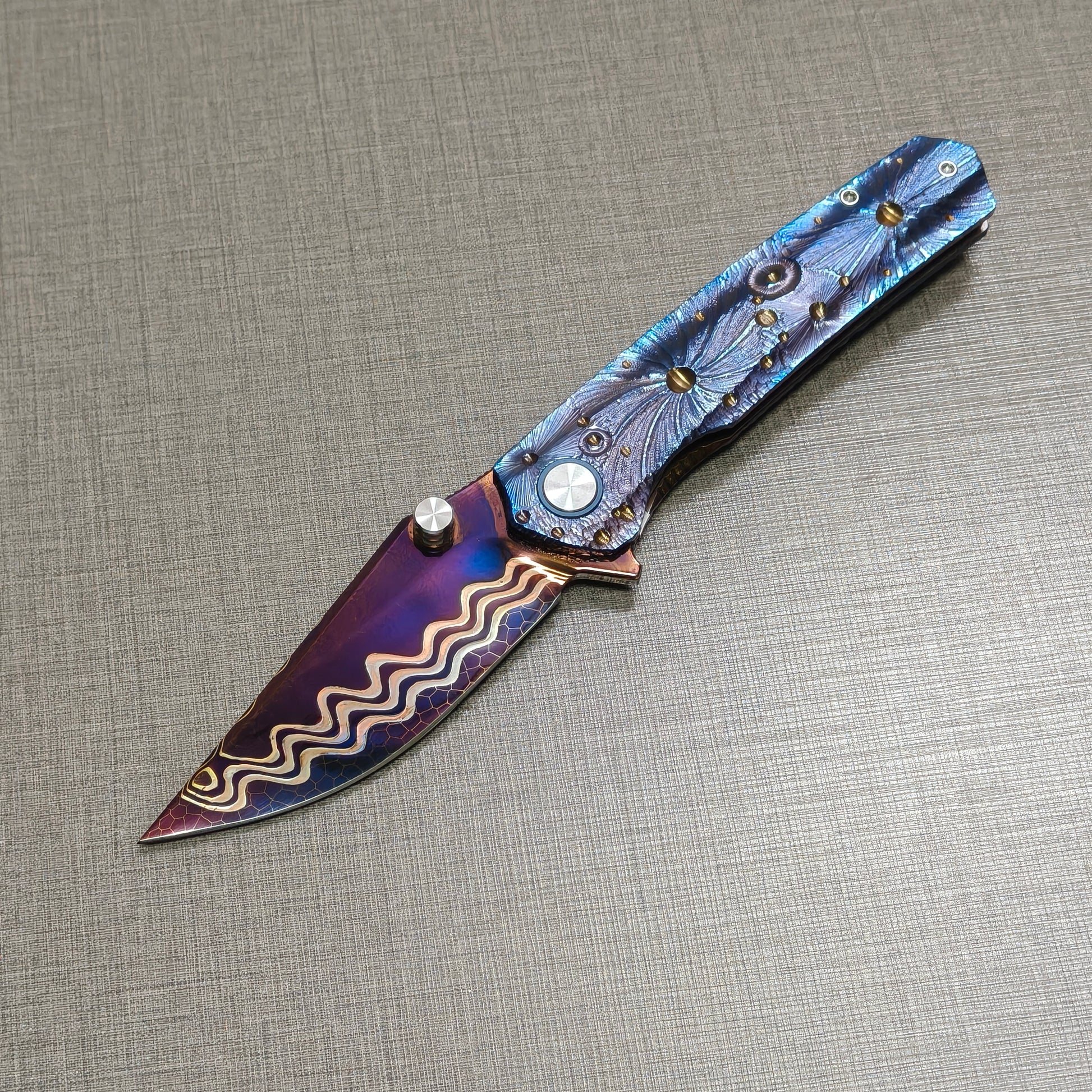 Impact Origin folding knife open, non-clip side showing engraved titanium handle inspired by meteor craters, hand-flamed damascus blade