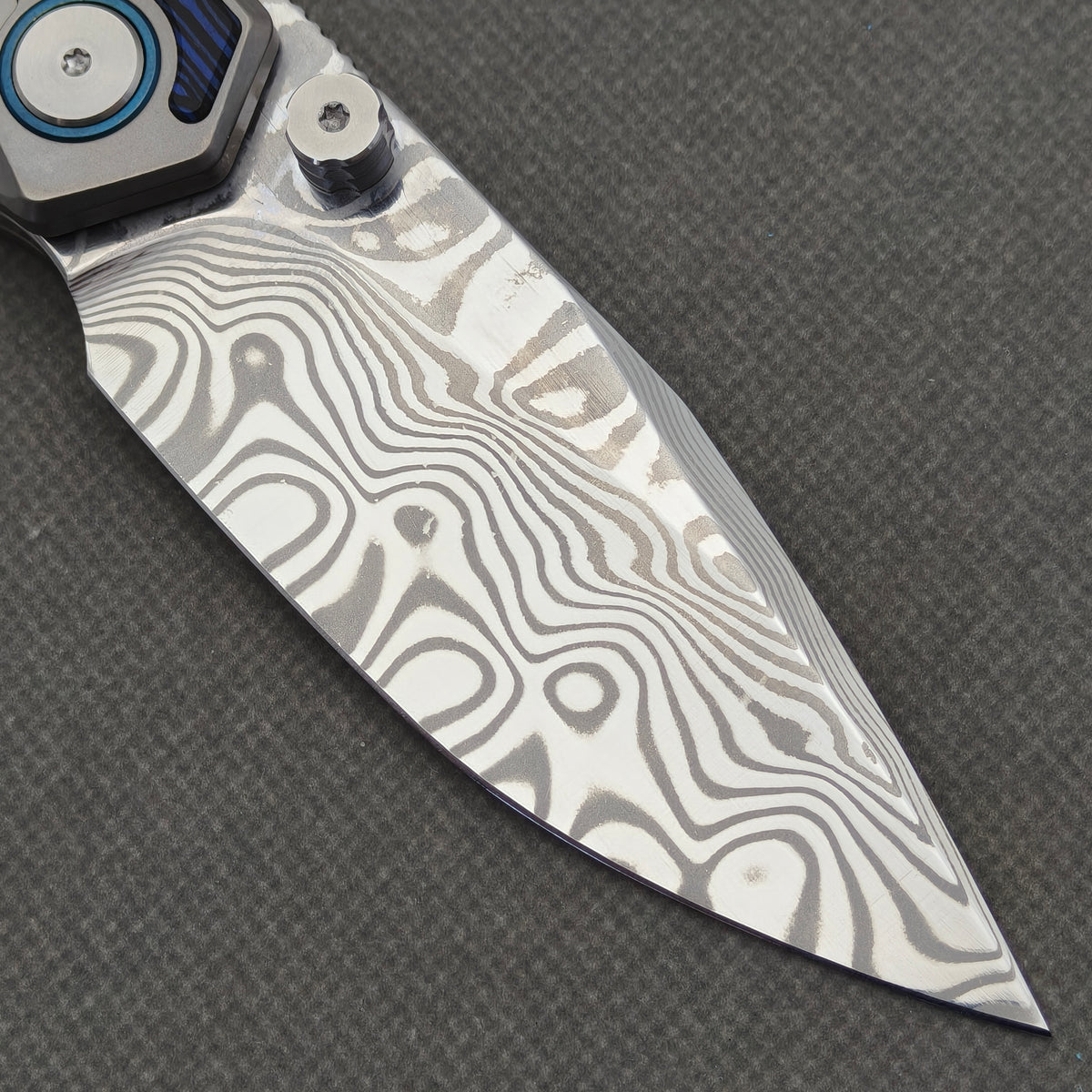 VORIXN Tidecipher KB157 Folding Knife – clip-side blade detail showing Damascus patterns, thumb stud, and blue pivot collar.