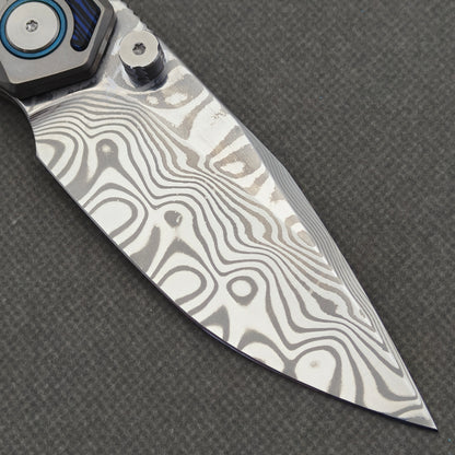 VORIXN Tidecipher KB157 Folding Knife – clip-side blade detail showing Damascus patterns, thumb stud, and blue pivot collar.